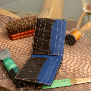 May include: Open bi-fold wallet with brown crocodile-embossed leather and blue pinstripe leather accents. The wallet has a brown stitched border. A leather brush, thread spool, and tools are in the background.