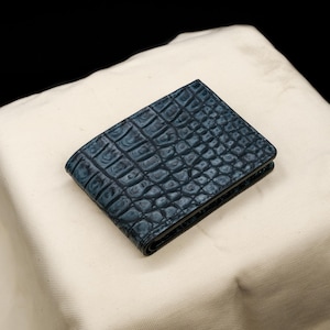 Puede incluir: Una cartera plegable de color azul oscuro con un patrón texturizado de piel de cocodrilo. La cartera tiene una forma rectangular con costuras visibles a lo largo de los bordes y un acabado brillante que resalta las escamas complejas.