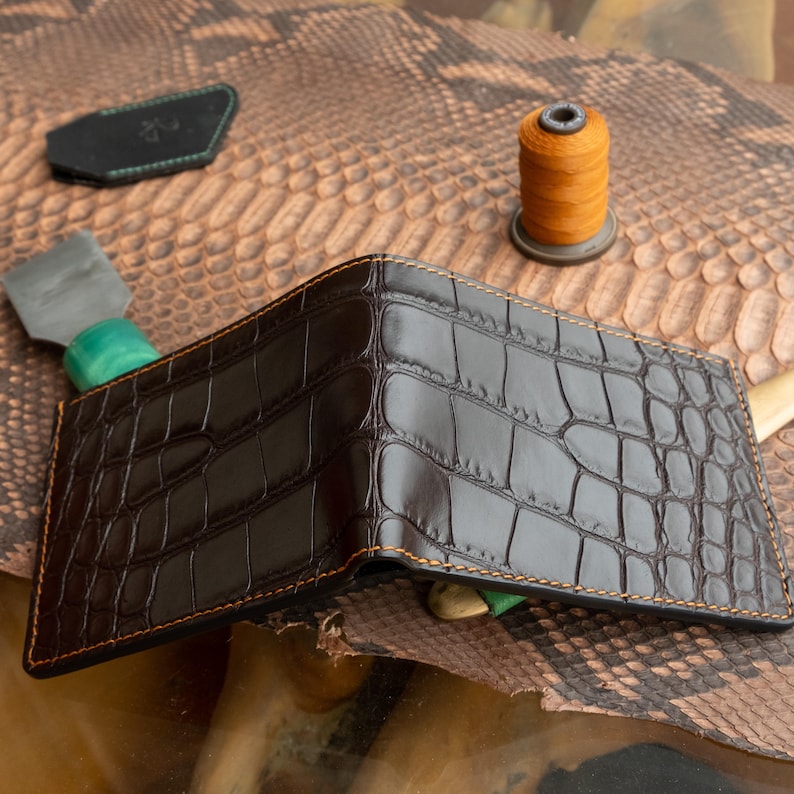 May include: Open brown leather wallet with a crocodile skin texture. The wallet has orange stitching around the edges. A spool of orange thread and a leather crafting tool are in the background.