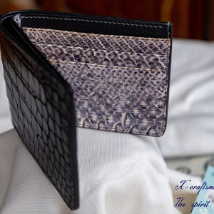 Puede incluir: Una cartera de cuero negra con un patr&oacute;n de cocodrilo en el exterior y un forro estampado en morado y blanco en el interior.