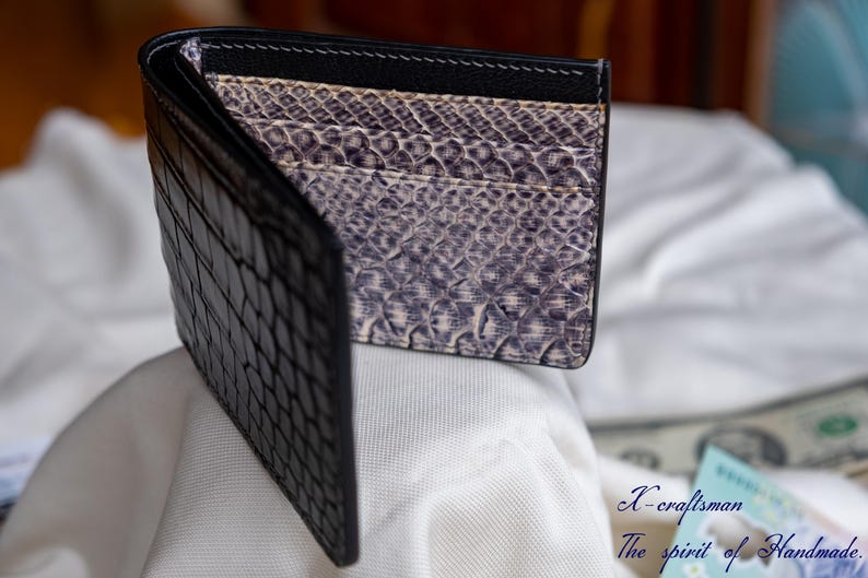Puede incluir: Una cartera de cuero negra con un patr&oacute;n de cocodrilo en el exterior y un forro estampado en morado y blanco en el interior.
