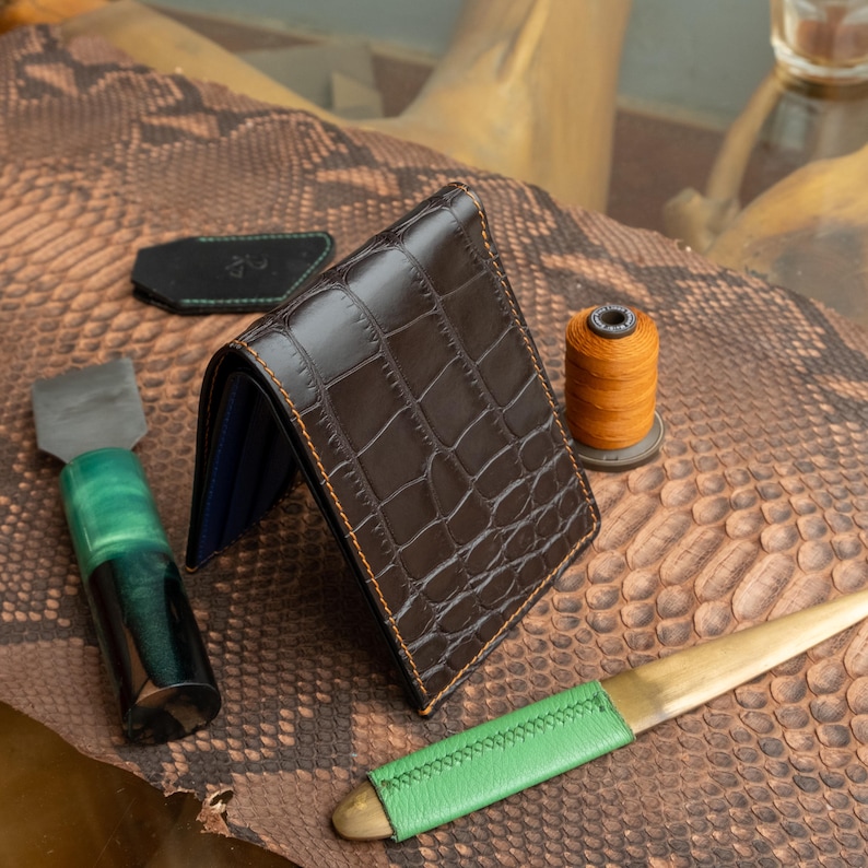May include: A brown crocodile-textured leather wallet with orange stitching, open to reveal a blue interior. A black leather tag with green stitching, a spool of orange thread, and a green-handled tool are also visible.