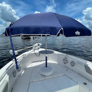 May include: A white boat with a blue and white umbrella mounted on the bow. The umbrella has a white trim and the words "The Ridge" printed on the canopy. The boat has a white table with cup holders and a blue base.