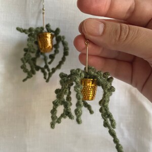 May include: A pair of gold-toned earrings with a green yarn plant hanging from each. The plant is in a small gold-toned thimble-shaped pot.