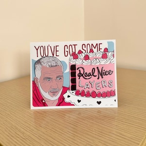 May include: A pink and white birthday card with a cartoon illustration of a man with a beard and a cake with pink frosting and strawberries. The text on the card reads "You've got some Real Nice Layers".