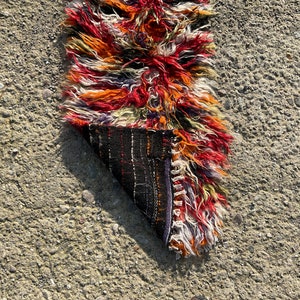 May include: A colourful, hand-woven rug with a long fringe. The rug is folded in half, revealing the underside, which is a dark brown with a geometric pattern.