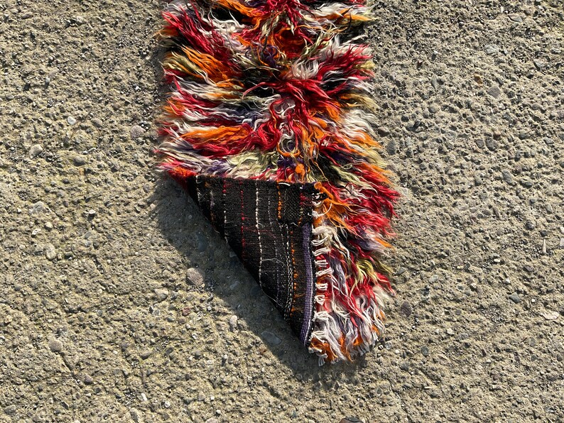 May include: A colourful, hand-woven rug with a long fringe. The rug is folded in half, revealing the underside, which is a dark brown with a geometric pattern.