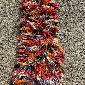 May include: A long, narrow rug with a shaggy texture. The rug is made of wool and has a colourful, abstract pattern in shades of red, orange, white, and purple.