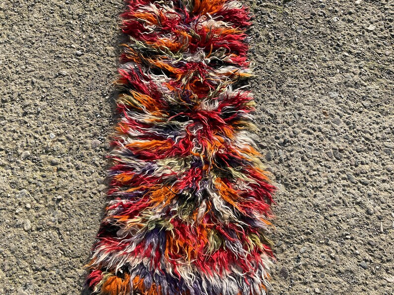 May include: A long, narrow rug with a shaggy texture. The rug is made of wool and has a colourful, abstract pattern in shades of red, orange, white, and purple.