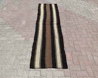 May include: A rectangular rug with vertical stripes in black, brown, and beige. The rug is made of a woven material and has a slightly frayed edge. The rug is placed on a brick surface.