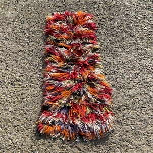 May include: A long, narrow, multi-coloured rug with a shaggy texture. The rug is made of wool and has a variety of colours, including red, orange, yellow, green, blue, and purple.