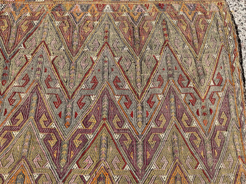 Pode incluir: Um close-up de um tapete tecido colorido com um padr&atilde;o geom&eacute;trico. O tapete apresenta um padr&atilde;o repetitivo de tri&acirc;ngulos e diamantes em tons de vermelho, laranja, amarelo, verde e azul.