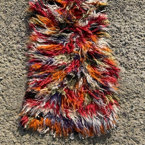 May include: A colourful, long, shaggy rug with a mix of red, orange, white, purple, and green wool fibres. The rug is lying on a grey, textured surface.