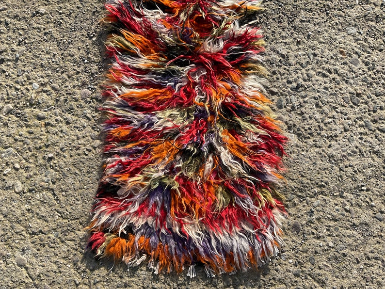 May include: A colourful, long, shaggy rug with a mix of red, orange, white, purple, and green wool fibres. The rug is lying on a grey, textured surface.