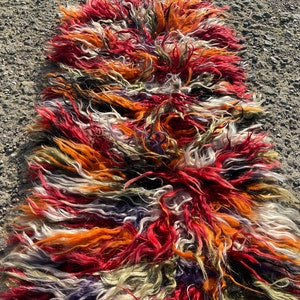 May include: A long, colourful rug made of shaggy, multi-coloured wool. The rug is a mix of red, orange, white, black, and green.