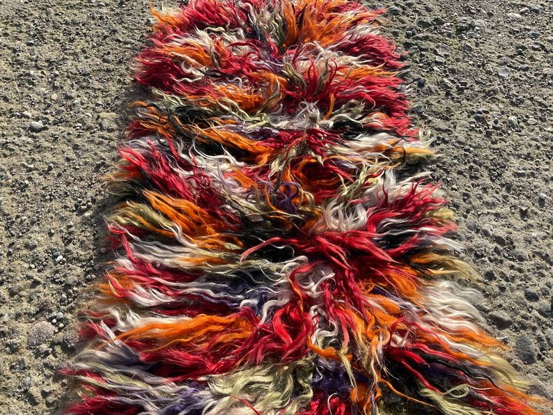 May include: A long, colourful rug made of shaggy, multi-coloured wool. The rug is a mix of red, orange, white, black, and green.