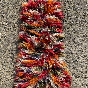 May include: A long, narrow rug with a shaggy texture. The rug is made of wool and has a colourful striped pattern in shades of red, orange, white, and black.