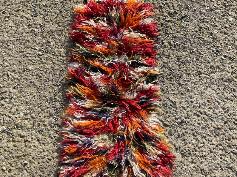 May include: A long, narrow rug with a shaggy texture. The rug is made of wool and has a colourful striped pattern in shades of red, orange, white, and black.