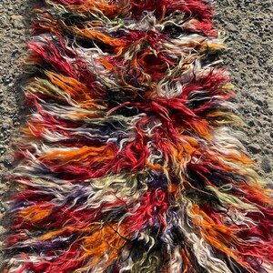 May include: A long, colourful rug made of shaggy, multi-coloured wool. The rug is made up of stripes of red, orange, white, black, and purple wool.