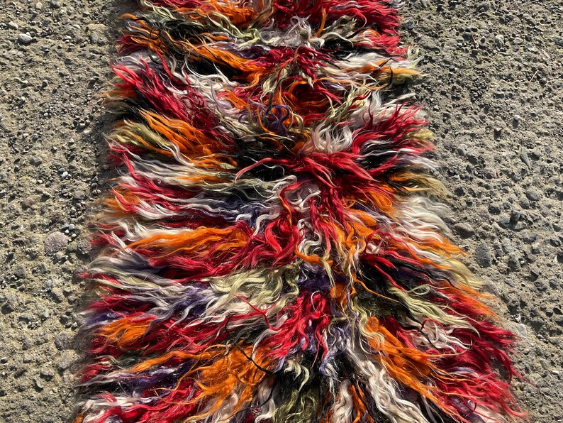 May include: A long, colourful rug made of shaggy, multi-coloured wool. The rug is made up of stripes of red, orange, white, black, and purple wool.