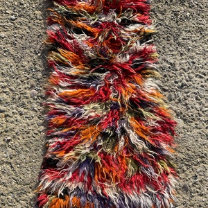 May include: A long, narrow rug with a shaggy texture. The rug is made of wool and has a colourful, abstract pattern in shades of red, orange, white, green, and purple.