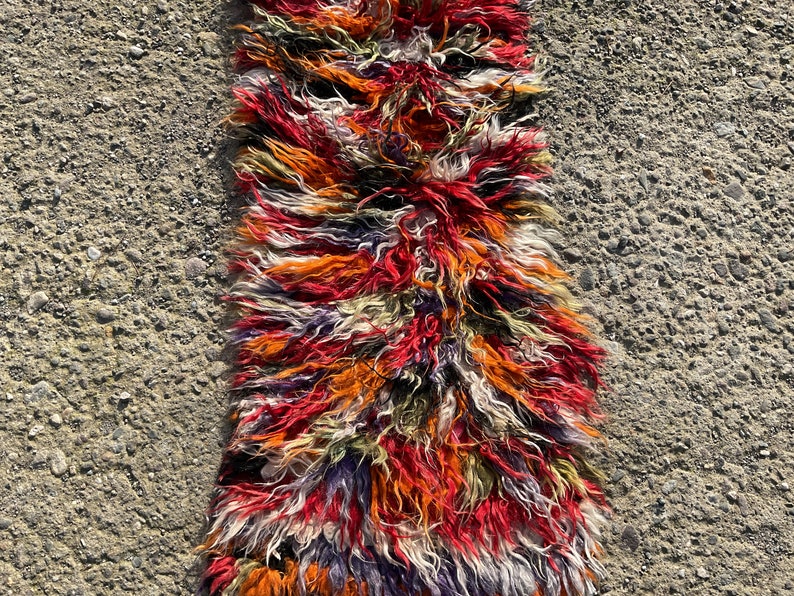 May include: A long, narrow rug with a shaggy texture. The rug is made of wool and has a colourful, abstract pattern in shades of red, orange, white, green, and purple.