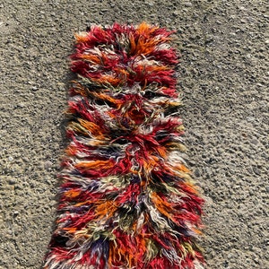 May include: A long, narrow rug with a shaggy texture. The rug is made of wool and has a colourful, abstract pattern in shades of red, orange, white, and black.