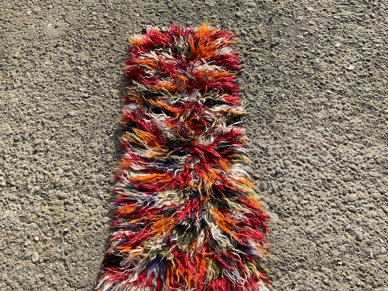May include: A long, narrow rug with a shaggy texture. The rug is made of wool and has a colourful, abstract pattern in shades of red, orange, white, and black.