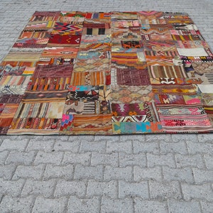 May include: A large, colorful patchwork rug made from various pieces of fabric. The rug is made up of many different colors and patterns, including red, blue, green, yellow, and brown. The rug is laid out on a gray concrete surface.
