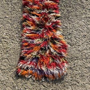 May include: A long, narrow rug with a shaggy texture. The rug is made of wool and has a colourful, abstract pattern in shades of red, orange, purple, and white.