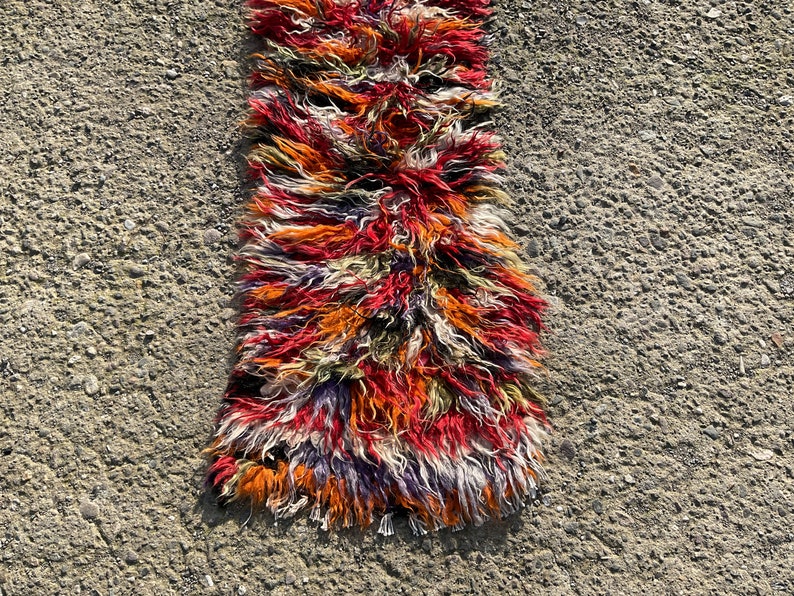 May include: A long, narrow rug with a shaggy texture. The rug is made of wool and has a colourful, abstract pattern in shades of red, orange, purple, and white.