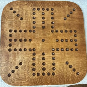 Puede incluir: Un tablero de juego de madera con una mancha marr&oacute;n. El tablero tiene un patr&oacute;n de cuadr&iacute;cula con 36 hendiduras circulares. Cada hendidura tiene una clavija marr&oacute;n oscura.