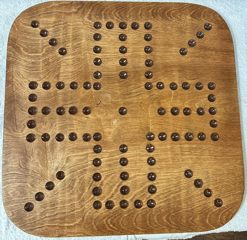 Puede incluir: Un tablero de juego de madera con una mancha marr&oacute;n. El tablero tiene un patr&oacute;n de cuadr&iacute;cula con 36 hendiduras circulares. Cada hendidura tiene una clavija marr&oacute;n oscura.