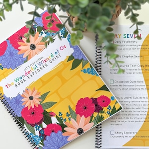 May include: A spiral-bound book titled "The Wonderful Wizard of Oz Book Explorer Guide" with a floral design. The cover features a yellow brick road background and colorful flowers. Another book with text is visible.