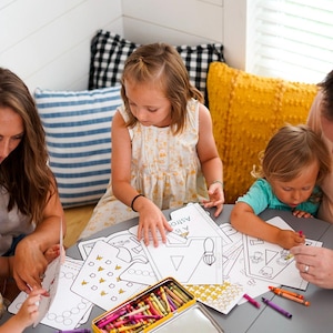 May include: A family of five sits around a round table, coloring. Coloring pages with geometric shapes and the word "Astronaut" are on the table. A metal crayon box and various crayons are also visible.