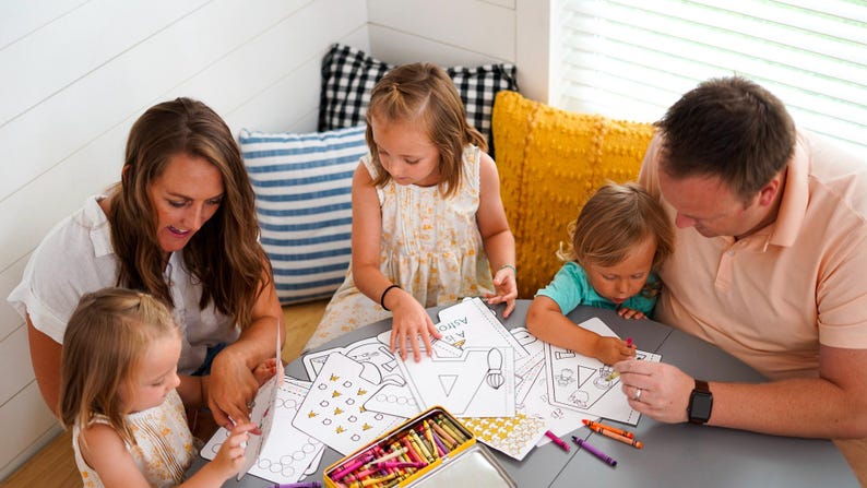 May include: A family of five sits around a round table, coloring. Coloring pages with geometric shapes and the word "Astronaut" are on the table. A metal crayon box and various crayons are also visible.