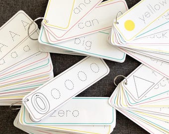 Puede incluir: Tarjetas didácticas blancas con esquinas redondeadas, con letras, números y formas. Cada tarjeta tiene un contorno punteado para trazar, con palabras como "amarillo" y "cero" impresas. Las tarjetas están unidas con anillas metálicas.