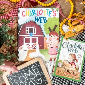 May include: Two copies of "Charlotte's Web" books, one spiral-bound and one paperback, with illustrations of farm animals. A small chalkboard with "SOME PIG" written on it is also present. The scene is decorated with yellow beads and red ribbon.