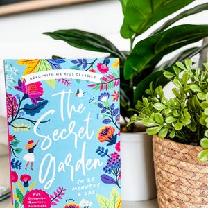 May include: A children's book titled "The Secret Garden" with a light blue cover and floral illustrations. The book is next to a potted plant with large green leaves and a small plant in a woven basket. The text on the book reads "Read-With-Me Kids Classics."