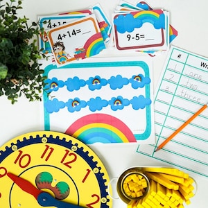 Op de afbeelding: Educatieve wiskundeset met regenboog-flashcards voor optellen en aftrekken, een werkblad met handgeschreven getallen, een gele klok met rode wijzers en een bakje met kleine gele telblokjes voor rekenoefeningen.