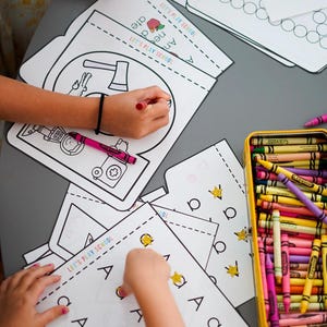 May include: Children's activity set with coloring pages and crayons. The pages feature illustrations and the text "LET'S PLAY SCHOOL". A yellow tin holds a variety of crayons. Gold star stickers are also present.