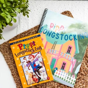 May include: Two children's books, "Pippi Longstocking" by Astrid Lindgren, are displayed on a woven mat. The book covers feature colorful illustrations of the character Pippi and her house. A potted green plant is in the upper left corner.