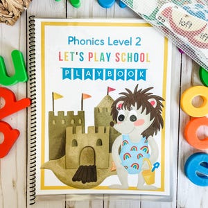 May include: A spiral-bound educational workbook titled "Phonics Level 2 LET'S PLAY SCHOOL PLAYBOOK" with a cartoon hedgehog in a swimsuit on the cover. Colorful magnetic letters and a mesh bag with word cards are scattered around the book.