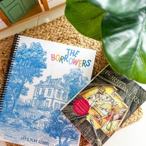 May include: Two books, "The Borrowers" and a spiral-bound notebook with a blue illustration of a house and trees. The notebook has the text "The Borrowers" in colorful letters and "Let's Play School". The books are on a woven mat.