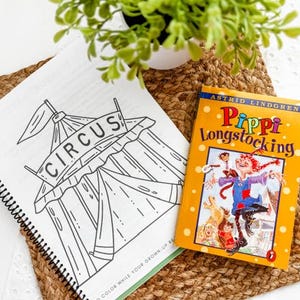 May include: A coloring book with a circus tent illustration and the word "CIRCUS" is open next to a copy of "Pippi Longstocking" by Astrid Lindgren. The books are on a woven mat with a small potted plant.