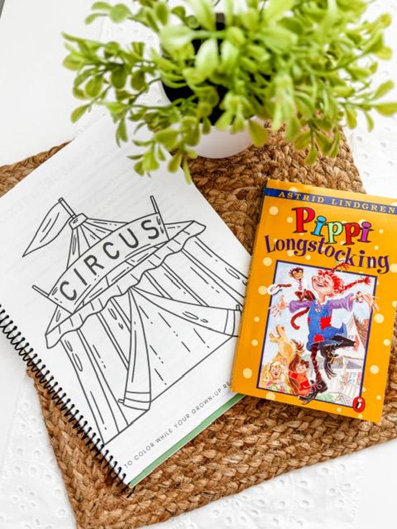 May include: A coloring book with a circus tent illustration and the word "CIRCUS" is open next to a copy of "Pippi Longstocking" by Astrid Lindgren. The books are on a woven mat with a small potted plant.