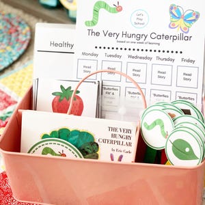 May include: A pink metal storage bin containing children's educational resources. It features a book titled "The Very Hungry Caterpillar", flashcards, and a weekly learning chart with a caterpillar and butterfly illustration.