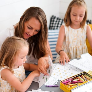 May include: A woman and two young children are seated at a gray table, engaged in an educational activity. They are using coloring pages and crayons from a yellow tin. The children are wearing yellow floral dresses.