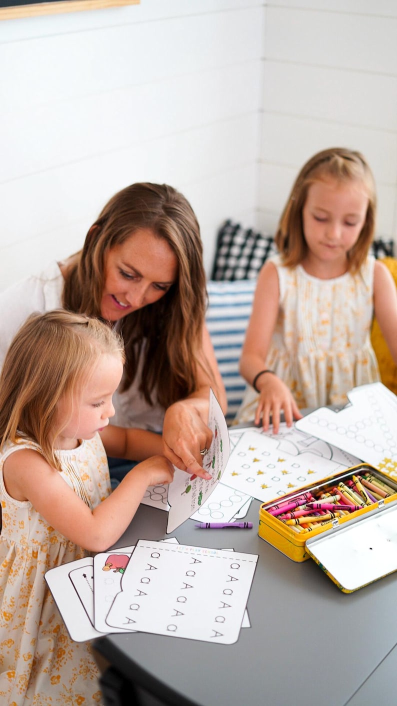 May include: A woman and two young children are seated at a gray table, engaged in an educational activity. They are using coloring pages and crayons from a yellow tin. The children are wearing yellow floral dresses.