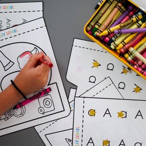 May include: A child coloring a worksheet with a red crayon. The worksheet has the text "LET'S PLAY SCHOOL" and illustrations of an apple, an axe, and a medical kit. A yellow crayon box filled with crayons is in the background.